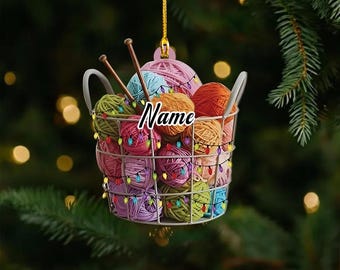May include: A decorative ornament shaped like a wire basket filled with colorful yarn balls and knitting needles. The ornament is adorned with string lights and the word "Name" in a cursive font. The background is a blurred image of a Christmas tree.