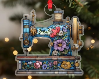 Puede incluir: Adorno de máquina de coser azul y dorado con detalles florales y el nombre "Sarah". El adorno tiene un efecto de vidriera, con una cinta roja para colgar. El diseño incluye una bobina de hilo y varias flores coloridas.