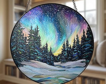 Op de afbeelding: Een ronde glas-in-loodpaneel met een winterlandschap en een levendige aurora borealis. De scène omvat besneeuwde grond, groenblijvende bomen en een kleurrijke lucht met tinten blauw, paars en geel. Een zwarte metalen ketting is bevestigd om op te hangen.
