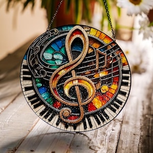 May include: A round stained glass suncatcher featuring a musical theme. The design includes a treble clef, musical notes, and a piano keyboard. The background is a mosaic of colorful glass pieces in shades of blue, green, yellow, and red. A chain is attached for hanging.