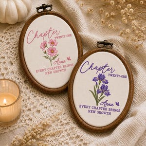 May include: Two oval wooden embroidery hoops containing white fabric with floral designs. One features pink flowers, the text "Chapter TWENTY-ONE", "Anna", and "EVERY CHAPTER BRINGS NEW GROWTH". The other has purple irises and identical text.