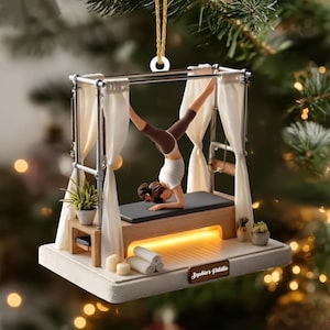May include: A Pilates-themed Christmas ornament. The ornament features a woman in a handstand on a Pilates reformer machine. The ornament has a white base with the text "Sophia's Pilates". The ornament is decorated with small plants, candles, and rolled towels.