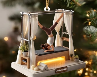 Peut inclure: Ornement de Noël sur le thème du Pilates. L'ornement représente une femme en équilibre sur les mains sur un appareil de Pilates. L'ornement a une base blanche avec le texte "Sophia's Pilates". Décoré de petites plantes, bougies et serviettes roulées.