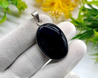 May include: A polished, oval-shaped black gemstone pendant with a silver-colored metal setting. The pendant is held against a white background with green and yellow foliage in the background. The pendant is approximately 1.5 inches long.