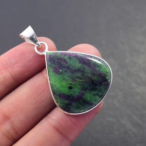 Peut inclure: Un pendentif en forme de goutte d'eau avec une pierre précieuse verte et violette sertie dans une lunette en argent. La pierre précieuse présente un motif naturel tourbillonnant. Le pendentif a une boucle en argent pour une chaîne.