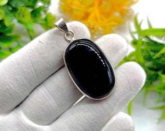 May include: A polished black oval pendant with a silver-colored metal frame and bail. The pendant is held against a white glove, with green foliage and yellow flowers in the background. The pendant is a jewelry item.