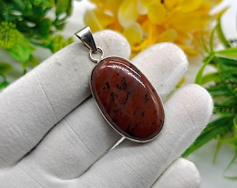 Pode incluir: Pingente oval com uma pedra polida castanho-avermelhada com inclusões pretas, emoldurada em uma armação de metal prateado. O pingente tem uma argola simples para uma corrente.