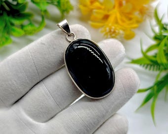 May include: Oval black gemstone pendant with a silver-colored metal frame and bail. The polished stone reflects light, and the pendant is set against a white background with green and yellow foliage.