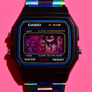 Könnte beinhalten: Eine digitale Casio F-91W Armbanduhr mit einem regenbogenfarbenen Metallarmband und Gehäuse. Das schwarze Zifferblatt hat blaue Akzente, den Text CASIO F-91W, ALARM CHRONOGRAPH, WATER WR RESIST und eine Digitalanzeige mit 22:34:32 und Grafik.