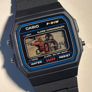 Könnte beinhalten: Schwarze Casio F-91W Digitaluhr mit schwarzem Armband. Das Zifferblatt hat einen blauen Rand und zeigt die Uhrzeit und ein kleines Bild an. Funktionen: Alarm, Chronograph und Wasserdichtigkeit. Klassisches Digitaldesign.
