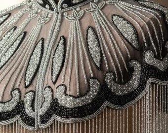 May include: A beaded cape displayed on a wooden mannequin. The cape features a high collar and a detailed design with black and silver beads. Long strands of silver beads cascade down, creating a fringe effect. The mannequin is on a wooden stand.
