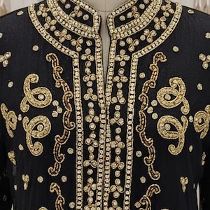 Peut inclure: Vêtement noir à manches longues avec broderies et ornements dorés complexes. Le design présente un col montant et des motifs ornés, notamment des formes de type cachemire et des rangées de petits accents scintillants. Le vêtement est présenté sur un mannequin dans une boutique.