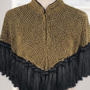 May include: A gold-coloured, textured cape with black fringe. The cape is displayed on a mannequin with a gold stand. The garment has a high collar and a central zip. The cape is a fashion accessory.