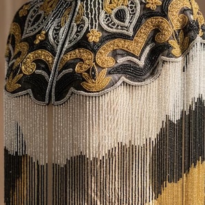 Puede incluir: Una capa con cuentas sobre un maniquí. La capa presenta intrincados bordados dorados, negros y plateados alrededor del cuello y los hombros. Largas franjas de cuentas doradas, negras y blancas cuelgan de la parte inferior. La capa se exhibe en un maniquí color crema.