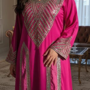 May include: A magenta-colored, long-sleeved dress with intricate silver embellishments around the neckline, sleeves, and down the front. The dress has a flowing silhouette and is worn by a person in a well-lit room with a chandelier.