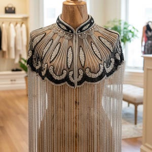 May include: A beaded cape on a wooden mannequin. The cape features a black collar and a detailed design with silver and white beads. Long silver fringe hangs from the bottom. The mannequin is on a metal stand in a well-lit room.