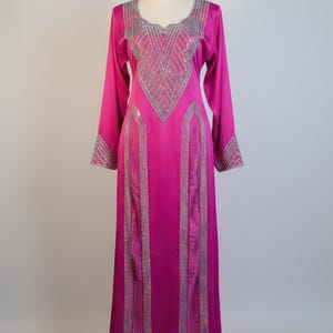 May include: A long, fuchsia-colored dress with long sleeves and a round neckline. The dress is embellished with silver-colored decorative accents on the neckline, sleeves, and front panels. The dress is displayed on a mannequin.