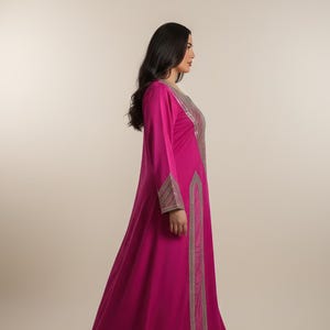 May include: A long, flowing magenta dress with long sleeves. The dress features intricate silver embroidery along the neckline, cuffs, and a vertical panel down the front. The dress is made of a soft, draping fabric.
