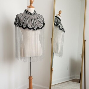 May include: A beaded cape with a high collar and silver fringe is displayed on a mannequin. The cape features a gray and black design. A gold-framed mirror reflects the cape and mannequin.
