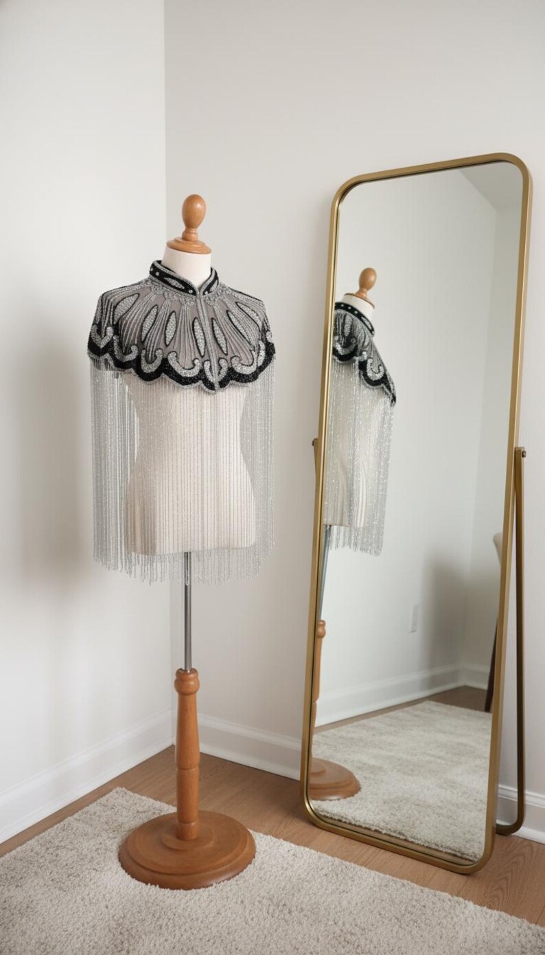 May include: A beaded cape with a high collar and silver fringe is displayed on a mannequin. The cape features a gray and black design. A gold-framed mirror reflects the cape and mannequin.