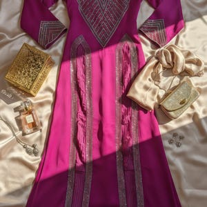 May include: A magenta-colored formal dress with long sleeves and intricate silver beaded embellishments. The dress features a V-neck design and vertical beaded stripes. Accessories include a gold clutch, scarf, and jewelry.