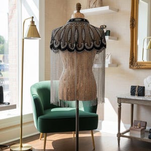 May include: A beaded cape with a black and silver design is displayed on a dress form. The cape features a scalloped edge and long, dangling silver fringe. The dress form is on a metal stand in a room with a green chair and gold lamp.