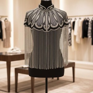 May include: A black mannequin displays a beaded cape with silver fringe. The cape features intricate silver and black beadwork around the neckline and shoulders, with long, vertical fringe. The background shows a clothing store.