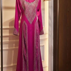 May include: A long, fuchsia-colored formal dress with long sleeves and a floor-length skirt. The dress features intricate silver embellishments around the neckline, cuffs, and down the front. It hangs on a wooden hanger.