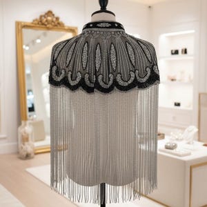 May include: A beaded cape with a high collar and long fringe. The cape is displayed on a mannequin. The collar and edges are black, with silver beading. The fringe is silver. The cape is in a boutique setting.