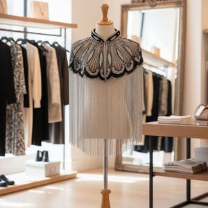May include: A beaded cape on a wooden mannequin. The cape features a black and silver design around the neckline and shoulders, with long silver fringe. The mannequin is in a boutique setting, with clothing racks and a mirror in the background.