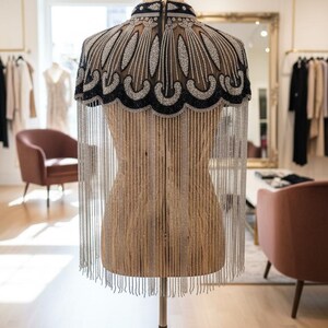 May include: A beaded cape displayed on a mannequin. The cape features a black collar and a scalloped design with silver and clear beads. Long silver fringe hangs from the bottom. The mannequin is in a boutique setting.