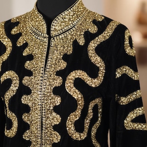 May include: Black velvet jacket with gold sequin embellishments. The jacket features a high collar and intricate, swirling patterns of sequins. The sequins are densely packed, creating a shimmering effect. The jacket is displayed on a black mannequin.