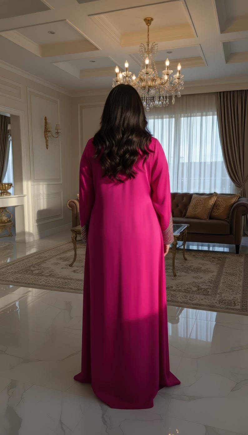 May include: A long, flowing magenta dress with long sleeves. The dress features intricate gold embroidery on the cuffs. The person is standing in a well-lit room with a chandelier and a patterned rug.
