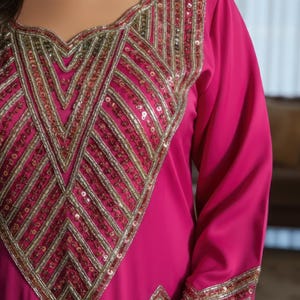 May include: A magenta-colored dress with long sleeves and intricate gold and sequin embellishments. The neckline and cuffs feature detailed geometric patterns. The dress has a flowing, elegant design, suitable for special occasions.