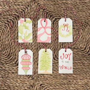 May include: Six watercolor Christmas gift tags with red and white twine. Designs include holly, a pink bow, a wreath, a red and green ornament, a Christmas tree, and the words "Joy to the World" in red.