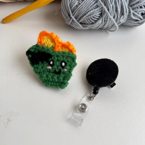 Könnte beinhalten: Ein handgefertigtes grünes Häkelabzeichen mit orangefarbenen und gelben Akzenten, das ein Cartoon-Gesicht und eine schwarze Augenklappe zeigt. Neben dem Häkelstück befindet sich eine schwarze, einziehbare Ausweisrolle. Häkelgarn und ein gelber Häkelhaken sind im Hintergrund.