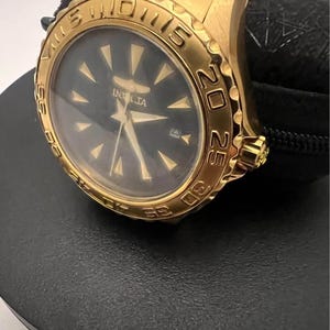May include: A gold-tone analog wristwatch featuring a black dial with gold-tone indices and hands. The watch has a rotating bezel with engraved numerals and a matching gold-tone metal link bracelet. The brand name Invicta is printed on the dial.