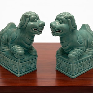 Puede incluir: Un par de estatuas de perros foo de cerámica verde celadón a juego. Cada criatura está representada en posición sentada sobre una base rectangular con un patrón geométrico de greca. Las figuras tienen crines estilizadas y bocas abiertas.