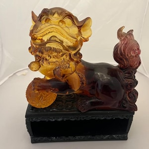 May include: A detailed amber-colored sculpture of a mythical lion-like creature, holding a ball. The sculpture has a dark base and a reddish-purple tail. The lion's mane and body are intricately detailed.