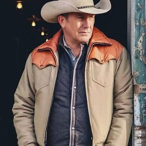Op de afbeelding: Een man draagt een beige cowboyhoed met een zwarte band. Hij draagt een beige jas met bruine schouderstukken, een donker gewatteerd vest en een blauw overhemd. De man leunt tegen een verweerde, groen geschilderde houten constructie.
