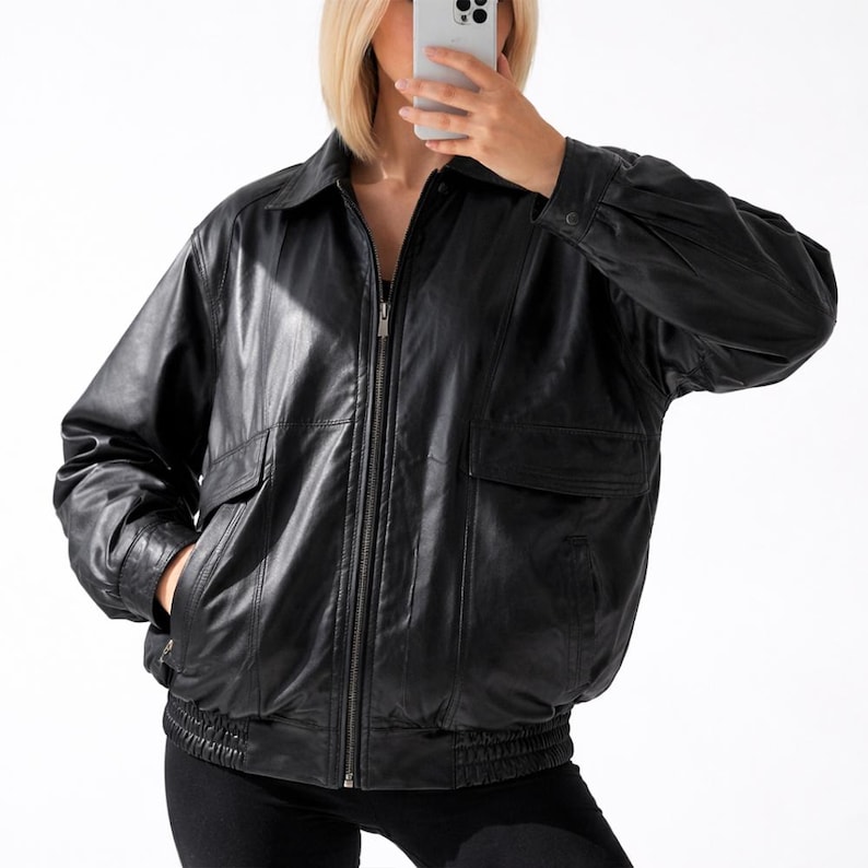 May include: Black leather jacket with a zip-up front, two front pockets, and a collared neckline. The jacket has a ribbed hem and cuffs. The sleeves are long and slightly puffed at the shoulders. The jacket is worn over a black top and black pants.