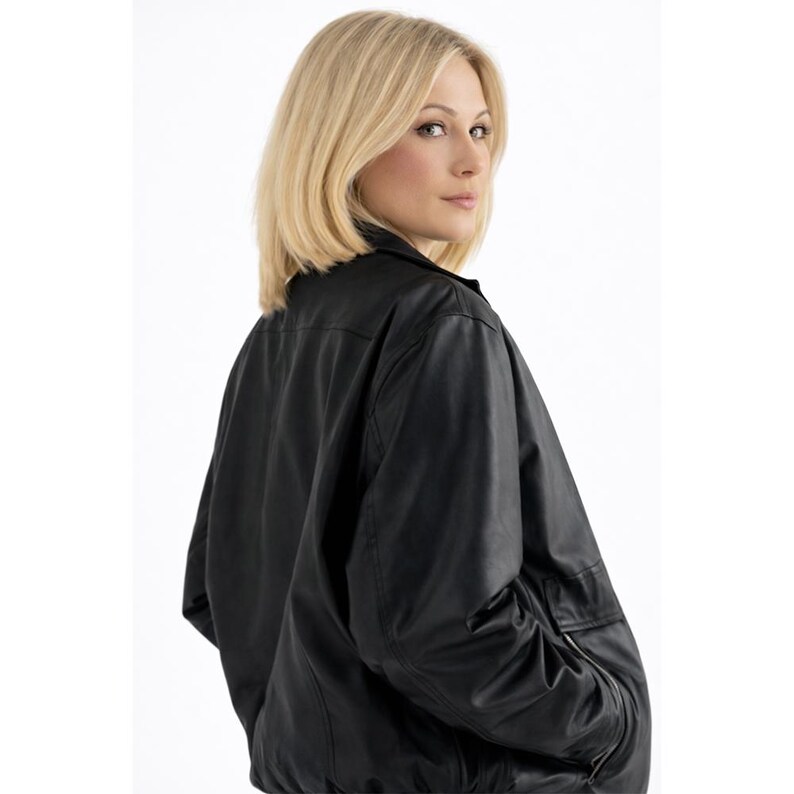 May include: A black leather jacket with a classic bomber style. The jacket features a collar, a front zipper, and a pocket on the left sleeve. The model has blonde hair and is looking over their shoulder.