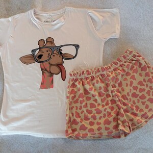 Puede incluir: Conjunto de pijama blanco con un gr&aacute;fico de jirafa con gafas y sacando la lengua. Los pantalones cortos tienen un fondo beige claro con manchas de jirafa rosas. La camisa tiene el texto "MAR & LEAF".