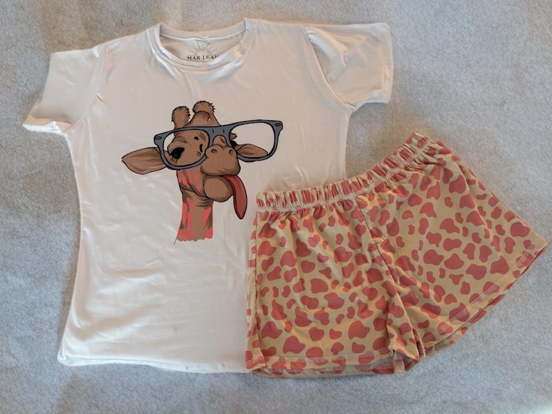Puede incluir: Conjunto de pijama blanco con un gr&aacute;fico de jirafa con gafas y sacando la lengua. Los pantalones cortos tienen un fondo beige claro con manchas de jirafa rosas. La camisa tiene el texto "MAR & LEAF".