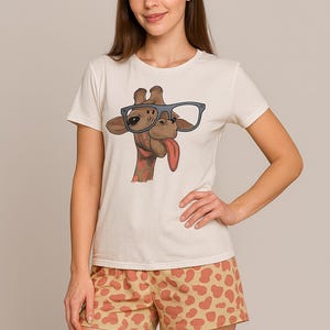 Puede incluir: Camiseta color crema con un dise&ntilde;o de jirafa con gafas y sacando la lengua. La jirafa es marr&oacute;n con la lengua roja. Los pantalones cortos son color beige con un patr&oacute;n naranja. El conjunto es informal.