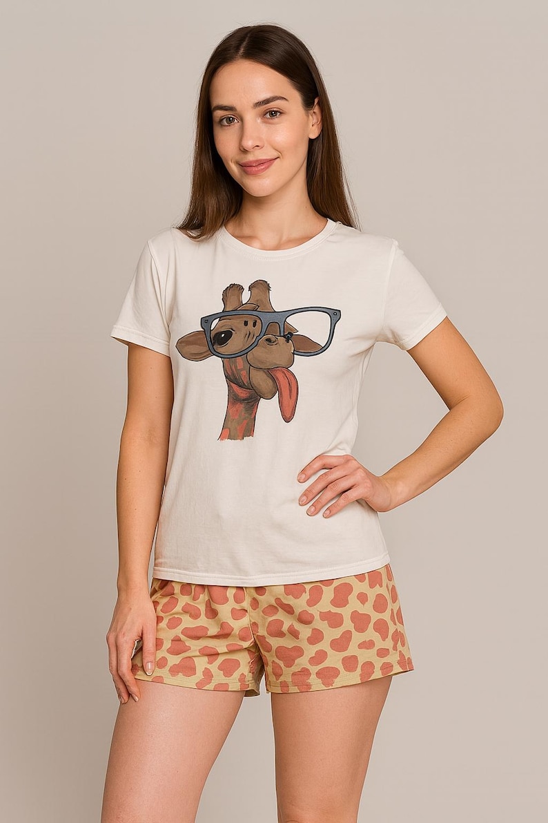 Puede incluir: Camiseta color crema con un dise&ntilde;o de jirafa con gafas y sacando la lengua. La jirafa es marr&oacute;n con la lengua roja. Los pantalones cortos son color beige con un patr&oacute;n naranja. El conjunto es informal.