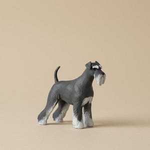 Puede incluir: Figura de un Schnauzer miniatura gris y blanco. El perro tiene una larga barba y cejas blancas, con detalles blancos en las patas y el pecho. La figura est&aacute; sobre una superficie de tono neutro.