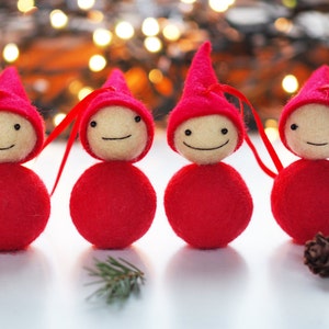 May include: Four handmade Christmas ornaments. Each ornament features a red body and pointed hat, with a cream-colored face and a stitched smile. Red ribbon loops are attached for hanging. The background includes blurred lights and pine needles.