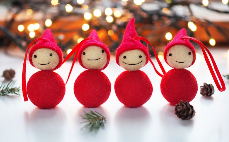 May include: Four handmade Christmas ornaments. Each ornament features a red body and pointed hat, with a cream-colored face and a stitched smile. Red ribbon loops are attached for hanging. The background includes blurred lights and pine needles.