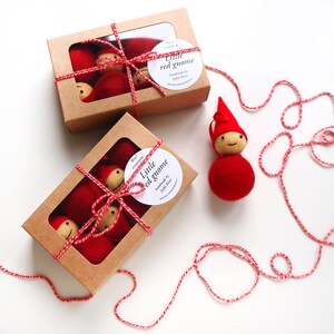 May include: Two cardboard boxes with clear windows, each containing several red gnome ornaments. The boxes are tied with red and white twine. A single gnome ornament sits beside the boxes. The ornaments have red bodies and hats, with beige faces. The text "Little red gnome" is on a white sticker.